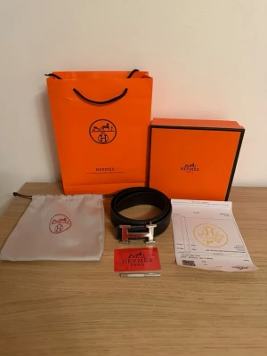 Svart bälte med H-spänne - Säljer ett svart bälte i skinn med det ikoniska H-spännet i silverfärg. Bältet har vit kontrastsöm och kommer med originalförpackning, dustbag och äkthetskort. Perfekt accessoar för att lyfta vilken outfit som helst. Storlek 110