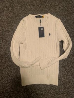 Vit kabelstickad tröja Polo Ralph Lauren - Kabelstickad vit tröja från Polo Ralph Lauren i bomull. Klassisk modell med rund hals, ribbade muddar och det ikoniska mörkblå broderade logot på bröstet. Perfekt till jeans och ger en stilren look. Pris ej hugget i sten.