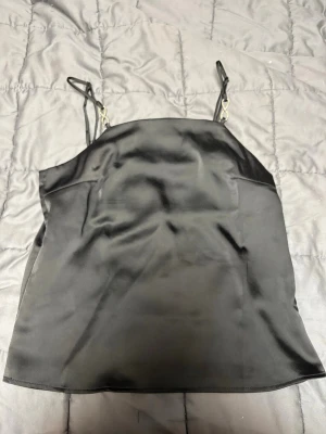 Svart satinlinne från H&M med strassdetalj - Säljer ett svart linne från H&M i satinliknande material. Linnet har tunna axelband med en snygg strassdetalj som ger extra bling. Modellen är rak och något lösare i passformen,  Storlek XS.