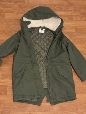 Grön parkajacka med huva från C - Säljer en grön parkajacka från C med mysigt foder i huvan och quiltat innerfoder. Jackan har dragkedja, tryckknappar och två stora fickor framtill. Perfekt för kalla dagar och har en klassisk, lite längre passform. Storlek 140- ålder 10