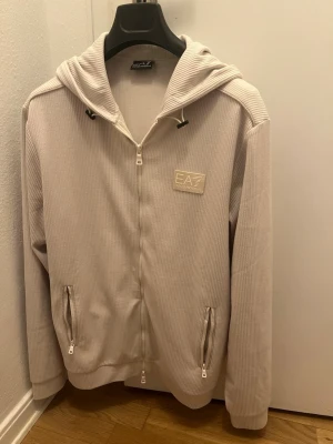 Emporio Armani zip hoodie - Stilren vit/ljus beigr EA7 zip hoodie med luva i väldigt skönt material med en logga som smälter in med hoodien på ett snyggt sätt. Storlek L. Mycket bra skick, har bara hängt i garderoben. Har ej sett denna hoodie i Sverige eftersom den är från London så den är rätt sällsynt. 