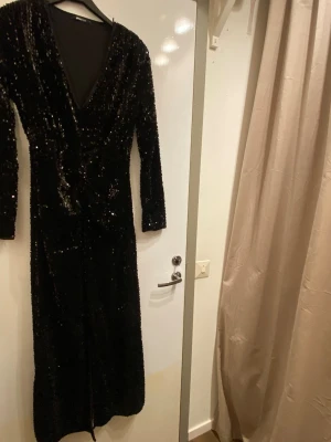 Svart glittrig långklänning Gina Tricot - Säljer en svart långklänning från Gina Tricot med glittriga paljetter över hela klänningen. Djup v-ringning, långa ärmar och en snygg slits framtill. Perfekt för dig som vill sticka ut på festen!