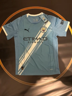Manchester City Haaland matchtröja - Manchester City matchtröja med Haaland 9 på ryggen. Tröjan är ljusblå med vita diagonala ränder, klubbmärke på bröstet och Etihad Airways som sponsor. Tillverkad i lätt polyester med vita detaljer vid ärmslut och hals. Perfekt för fotbollsfans!