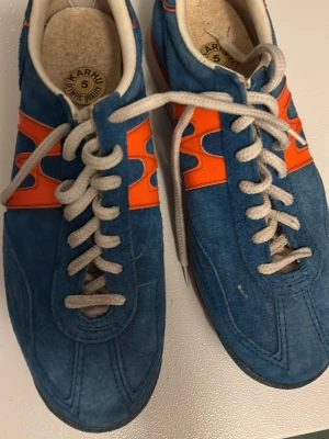 Blå-orange retro sneakers från Karhu - Säljer ett par retro Karhu sneakers i blå mocka med orange detaljer och vita snören. Skorna har en klassisk låg profil och rund tå, perfekt för dig som gillar vintagevibbar och sportig stil. Karhu-logga på plösen och sidorna.