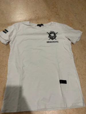 Vit Ibrahimovic t-shirt Concrete Humanity - Snygg vit t-shirt från Concrete Humanity med Ibrahimovic-tryck och nummer 10 på ryggen. Svensk flagga på ärmen och coolt emblem på bröstet. Skön passform och tillverkad i mjuk bomull. Perfekt för dig som gillar sportig stil och vill sticka ut.