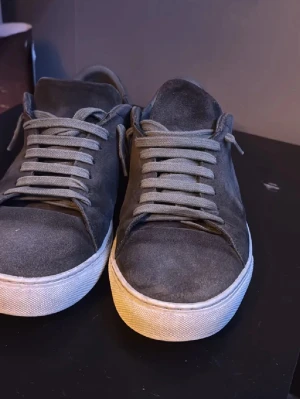 Grå mocka sneakers från Axel Arigato - Snygga sneakers från Axel Arigato i mörkgrå mocka med vita, tjocka gummisulor och matchande grå snörning. Skorna har en stilren design med rund tå och diskret logga på hälen och sulan. Perfekta för dig som gillar minimalistisk och clean stil. Pris kan diskuteras vid snabb affär!