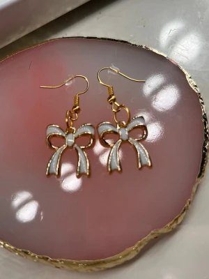 Elfenbensglans💍 - Säljer ett par söta örhängen formade som rosetter i vit emalj med guldfärgade kanter. De har krokfäste och är perfekta för dig som gillar detaljer med lite extra sötma. Passar till många olika stilar och ger en lekfull touch till din outfit💎