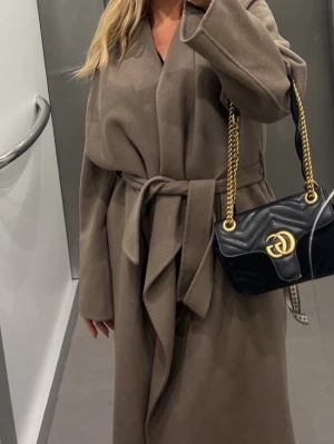 Beige omlottkappa med bälte Ivy oak  storlek 36 - Säljer en snygg beige kappa i omlottmodell med bälte i midjan. Jackan har långa ärmar och en stilren, enkel design som ger en chill och trendig vibe. Perfekt för dig som gillar klassiska plagg med modern touch. Ull nypris 5000