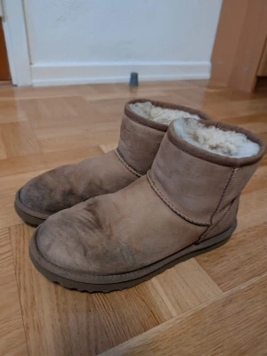 Bruna UGG boots i mocka - UGG boots i klassisk modell med brun mocka på utsidan och mjukt, ljust foder av fårskinn på insidan. Skorna har rund tå, platt sula och en enkel design utan snörning. Perfekta för kalla dagar och ger en cozy vibe.