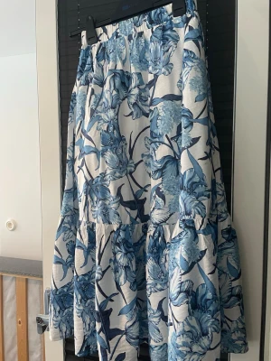 Blommig långkjol från Dobber XS - Supersnygg långkjol från Dober i storlek XS. Kjolen har ett vitt tyg med blått blommönster och bred resår i midjan för skön passform. Den har volangdetaljer och är perfekt för sommarens alla dagar. Materialet känns lätt och luftigt. Har använts 2 ggr.