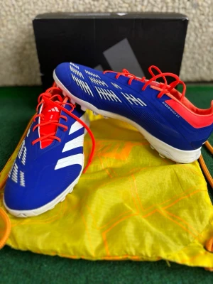 Adidas Predator blå fotbollsskor - Säljer ett par Adidas Predator fotbollsskor i blått med vita detaljer och röda snören. Skorna har en låg profil, rund tå och platt sula med dobbar för bra grepp på konstgräs. Ovandelen är i syntetmaterial och insidan har en röd kontrast. Perfekt för dig som vill sticka ut på planen.  Skickar även med en gratis påse som du ser på bild 2. Den är inte så mycket använt, men bara att konstgräs färgen stanna kvar. Den är för Konstgräs,Turf, utomhus skor. 