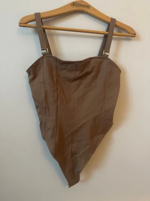 Brun croppad väst med breda axelband - Säljer en brun croppad väst med breda axelband och markerade sömmar. Västen har en spetsig nederkant och är tillverkad i ett mjukt material som ger en snygg siluett. Perfekt att styla över en t-shirt eller skjorta för en trendig look.