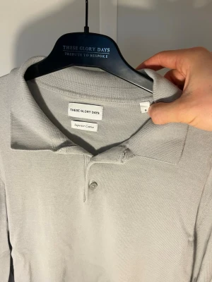Ljusgrå långärmad piké från These Glory Days - Säljer en stilren ljusgrå pikétröja från These Glory Days i mjuk bomull. Tröjan har klassisk krage, knäppning med tre knappar och långa ärmar. Det är kameran och ljuset som gör de ser skumt på bild 3.