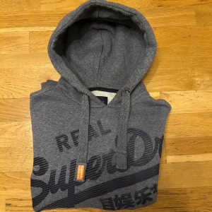 Superdry hoodie  - Säljer en grå hoodie från SuperDry med svart tryck på framsidan och en grön detalj på ärmen. Den har en klassisk huva med snörning och ribbade muddar vid ärmslut och nederkant. Perfekt för en avslappnad stil.