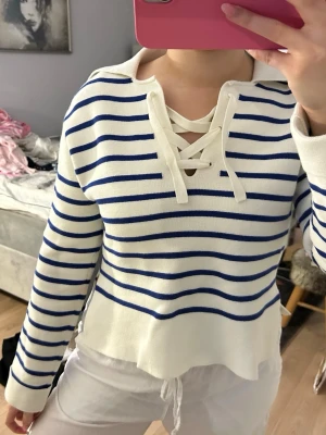 Randig vit-blå stickad tröja med snörning - Säljer en vit stickad tröja med blå ränder och snygg snörning vid halsen. Tröjan har lång ärm och en avslappnad passform, perfekt för vår eller sommarkvällar. Klassisk marint inspirerad look som är enkel att styla.