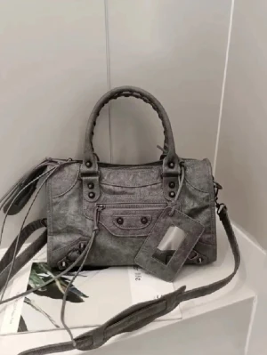 Grå axelväska med detaljer - Snygg grå axelväska i skinn med både handtag och justerbar axelrem. Väskan har coola metalldetaljer, dragkedja framtill och en praktisk tagg för kort. Perfekt för dig som vill ha en stilren men edgy look.