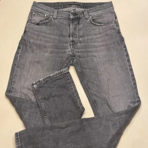 Grå jeans från Nudie Jeans - Säljer ett par grå jeans från Nudie Jeans med klassisk femficksdesign och svarta broderade detaljer på bakfickorna. Jeansen har en rak passform och är tillverkade i slitstarkt denimtyg. Snygg tvättad look och svart patch bak i midjan. Har tyvärr behövt laga jeansen men funkar bra, se på sista bilden. 