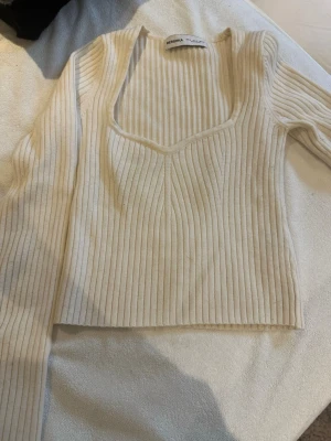 Ribbad ljus beige topp från Bershka - Säljer en ribbad beige topp från Bershka med lång ärm och djup v-ringning. Toppen är croppad och har en snygg, figurnära passform som framhäver siluetten. Perfekt att styla med höga jeans eller kjol.