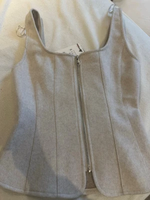 Grå korsettopp med dragkedja - Säljer en grå korsettopp från Zara i mjukt material. Toppen har markerade sömmar och en snygg dragkedja framtill som ger en cool look. Passformen är figurnära och toppen har breda axelband. Perfekt att styla med jeans eller kjol.