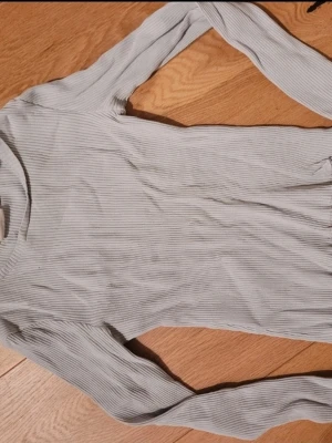 Ljusgrå ribbad långärmad topp - En stilren ljusgrå långärmad topp med smala ribbade ränder. Toppen har en enkel, kroppsnära passform och är tillverkad i ett mjukt och stretchigt material som känns skönt mot huden. Perfekt att matcha med jeans eller kjol för en clean look.