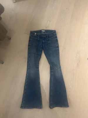 Blå bootcut jeans med slitningar - Säljer ett par blå bootcut jeans med snygga slitningar vid knäna och framfickor. Jeansen har låg midja och är tillverkade i klassiskt denimtyg. ❤️