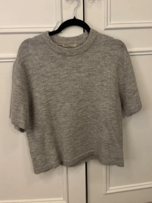 Grå stickad kortärmad tröja Gina Tricot - Mysig grå stickad tröja från Gina Tricot i kortärmad modell. Tröjan har rund halsringning och ribbade kanter för en clean look. Perfekt till lager på lager under höst och vår. Enkel att matcha med både jeans och kjol.