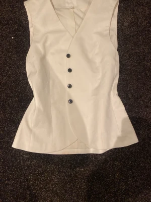 Vit figursydd väst med knappar - Säljer en stilren vit väst med figurnära passform och fyra svarta knappar framtill. Västen har v-ringning och är tillverkad i ett slätt tyg som ger en clean look. Perfekt att styla över en skjorta eller t-shirt för en trendig outfit.