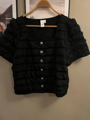 Svart blus med puffärm och knappar - Säljer en svart blus från Yas med korta puffärmar och dekorativa silverfärgade knappar framtill. Blusen har ett ribbat och volangigt tyg . Fyrkantig ringning 