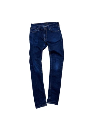 Nudie Jeans Skinny Lin blå jeans - Säljer ett par mörkblå Skinny Lin jeans från Nudie Jeans i storlek W28 L32. Modellen har klassisk femficksdesign, kontrastsömmar och Nudie-logga på bakfickan. Tillverkade i 99% ekologisk bomull och 1% elastan för stretchig passform.