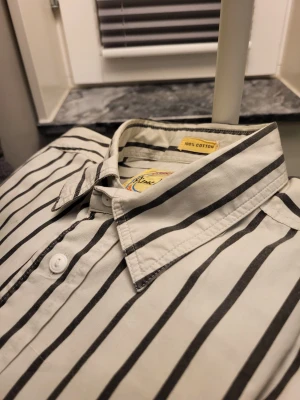 Vintage Randig skjorta från Pierre Hector - Snygg vit skjorta med svarta vertikala ränder från Pierre Hector Young Collection. Skjortan har klassisk krage, bröstficka med broderad krona i rött, blått och gult, samt vita knappar. Tillverkad i 100% bomull för skön känsla och bra passform.