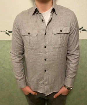 Grå overshirt från Riley - Snygg grå overshirt från Riley i bomull med klassisk krage och två bröstfickor med knappar. Skjortan har lång ärm och mörka knappar framtill. Passformen är slim och materialet känns mjukt och bekvämt. Perfekt för dig som gillar stilrena och enkla plagg.