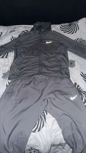 2 Nike grå träningssets med huva (säljs ej separat) - Nike träningsset i grått med jacka och byxa. Jackan har huva, dragkedja och två fickor, samt Nike-logga på bröstet. Byxan har elastisk midja och Nike-logga på benet. båda sets är storlek S och säljs tillsammans!