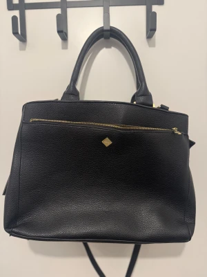 Svart handväska från Rizzo - Elegant svart handväska från Rizzo i strukturerat skinn, smyckad med guldfärgade detaljer och stilrena dragkedjor. Väskan är smart designad med flera praktiska fack både invändigt och utvändigt, inklusive ett särskilt laptopfack som rymmer upp till 15 tum. Rejäla och bekväma handtag gör den perfekt för både vardag och resa. Lätt slitage på handtagen, men i övrigt i mycket fint skick. På baksidan finns dessutom ett smidigt hölje som gör att du enkelt kan fästa väskan på resväskans handtag – perfe