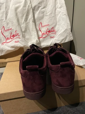Vinröda Christian Louboutin sneakers - Säljer ett par vinröda sneakers från Christian Louboutin i mjuk mocka med snörning och rund tå. Skorna har coola nitar på tån för extra edge och kommer med originalpåse och låda. Perfekt för dig som vill sticka ut med lyxiga detaljer. Är köpte i Italien/rom kvitto kommer även med ifall jag hittar det man kan även skicka dom till närmaste butik ifall man tappar nitar. Är bra skick har bara används en gång.
