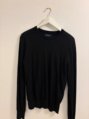 Svart merinoulltröja från Riley - Svart långärmad tröja från Riley i mjuk merinoull. Klassisk rund halsringning och ribbade muddar vid ärmslut och nederkant. Perfekt för dig som gillar stilrena och enkla plagg med skön passform. Knappt använd, Nypris 900kr på Brothers.