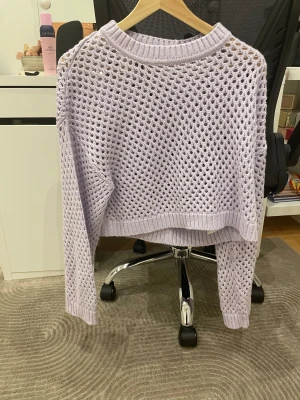 Ljuslila stickad croppad tröja - Säljer en ljuslila stickad tröja med hålmönster och croppad passform. Tröjan har långa ärmar och ribbade muddar vid hals, ärmslut och nederkant. Perfekt för dig som gillar en chill och trendig stil.