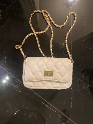 Vit quiltad axelväska med kedja - Snygg vit axelväska med quiltat mönster och guldfärgad kedja som axelrem. Väskan har en rektangulär form och ett elegant vridlås i guld på framsidan. Materialet ser ut som skinn och ger en lyxig känsla. Perfekt för att lyfta din outfit.