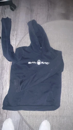 Svart Sail Racing hoodie med huva - Svart hoodie från Sail Racing med vit logga tryckt på bröstet. Tröjan har huva, lång ärm och en stor magficka framtill. Perfekt för dig som gillar sportiga och stilrena plagg. Materialet känns mjukt och bekvämt, troligen bomull eller polyester.