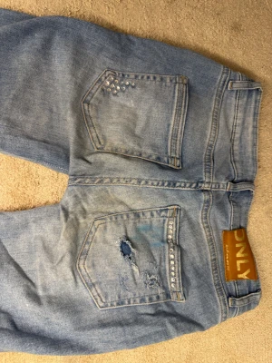 Bootcut jeans med pärldetaljer från ONLY - Bootcut jeans i ljusblå tvätt från ONLY, med låg midja och pärldetaljer på fickorna. Jeansen har slitningar och hål på ena bakfickan för en edgy look. Klassisk femficksmodell i mjukt denimtyg, perfekt för dig som gillar lite extra detaljer.