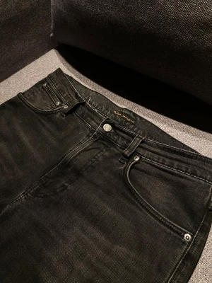 Svarta jeans från Nudie Jeans Co - Snygga svarta jeans från Nudie Jeans Co med klassisk femficksdesign och subtil broderad våg på bakfickan. Jeansen har raka ben och normal passform, tillverkade i mjukt denimtyg med metallknappar och nitar. Perfekt för dig som gillar stilrena och tidlösa jeans. Modellen är lean dean. Storlek 31/32. Skick 8/10