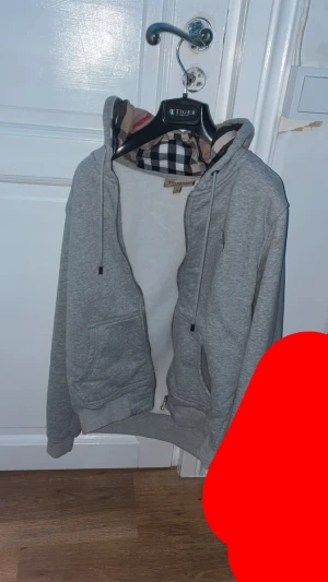 Grå Burberry hoodie med dragkedja - Grå hoodie från Burberry med klassiskt rutigt foder i huvan och insidan. Tröjan har snörning vid huvan, dragkedja framtill och två fickor. Långärmad och med ribbade muddar. Perfekt för dig som gillar stilren design med lyxig touch. Passar S också