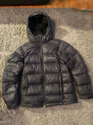 Svart Peak Performance dunjacka - Säljer en svart dunjacka från Peak Performance med huva och dragkedja framtill Jackan ny pris 3299kr, men jackan har ett håll i sig därför säljer jag den för 299kr. Jackan har ett quiltat mönster och är tillverkad i syntetmaterial med dunfyllning. Perfekt för kalla dagar och har en clean, sportig look. Hållet är inte synligt men det finns.