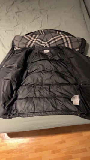 Svart pufferjacka från Burberry M - Säljer en svart pufferjacka från Burberry London England i storlek M. Jackan har en klassisk Burberry-rutig insida på huvan, stor huva och tydlig logga på ärmen. Materialet är polyester och jackan har dragkedja framtill. Perfekt för kalla dagar.                                                                                             Man är även justerbar att göra till en väst genom plocka bort armarna!     Pris kan diskuteras🤭