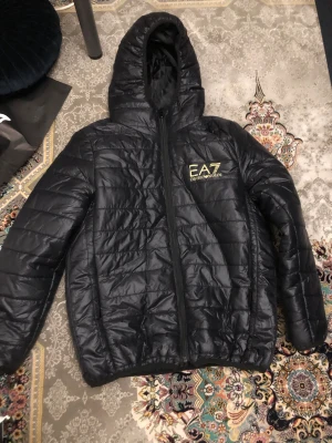 Svart pufferjacka EA7 Emporio Armani - Svart pufferjacka från EA7 Emporio Armani med huva och gul logga på bröstet. Jackan har quiltad design, dragkedja framtill och långärmade ärmar. Perfekt för dig som gillar stilren och sportig look. Materialet är syntet och jackan är lätt vadderad.
