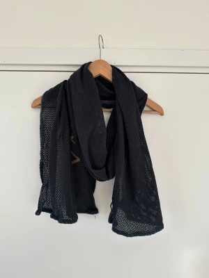 Svart halsduk från Vaillent - Svart halsduk från Vaillent med guldigt tryck längs ena sidan. Rektangulär form och lätt, luftig känsla. Perfekt att slänga över axlarna för en cool vibe. Materialet är syntet och ger ett sportigt uttryck.