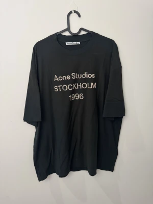 Svart Acne Studios t-shirt Stockholm unisex M relaxed fit - Svart t-shirt från Acne Studios med tryck 'Acne Studios Stockholm 1996' i vitt framtill. Modellen är loose fit med korta ärmar och rund hals. Materialet är mjuk bomull som känns skön mot huden. Perfekt för dig som gillar stilrena och coola statement-plagg.