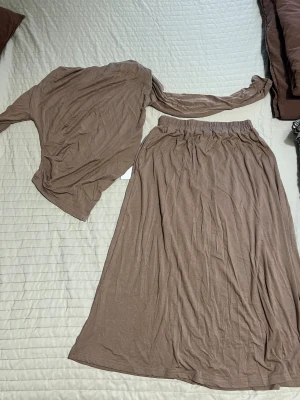 Brun långkjol i mjukt material - Säljer en enkel och flowy brun långkjol plus topp med resår i midjan. Kjolen är tillverkad i ett tunt och mjukt tyg som ger en avslappnad känsla. Perfekt för dig som gillar en stilren och bekväm look.