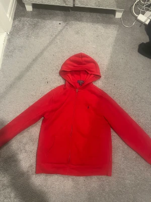 Röd hoodie från Polo Ralph Lauren - Säljer en röd hoodie från Polo Ralph Lauren i storlek XL. Tröjan har huva, dragkedja framtill och två fickor. Klassisk design med liten broderad logga på bröstet. Perfekt för chill dagar och enkel att matcha med jeans eller joggers.