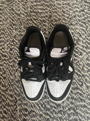 Nike Dunk Low svart/vit sneakers - Nike Dunk Low sneakers i svart och vitt med klassisk Swoosh på sidan. Skorna har snörning, perforerad tå och platt sula. Tillverkade i skinn med textilfoder. Stilren och tidlös modell som är populär bland sneakerheads.