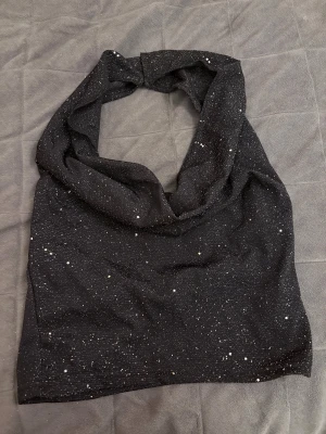 Svart glittrig halterneck topp - Säljer en svart halterneck topp med glittrigt mönster. Toppen är ärmlös och har en snygg knäppning i nacken. Materialet är mjukt och stretchigt syntet, perfekt för att sticka ut med lite extra shine. Tyget är dock väldigt tunt men är inte genomskinligt. Har endast testat toppen.